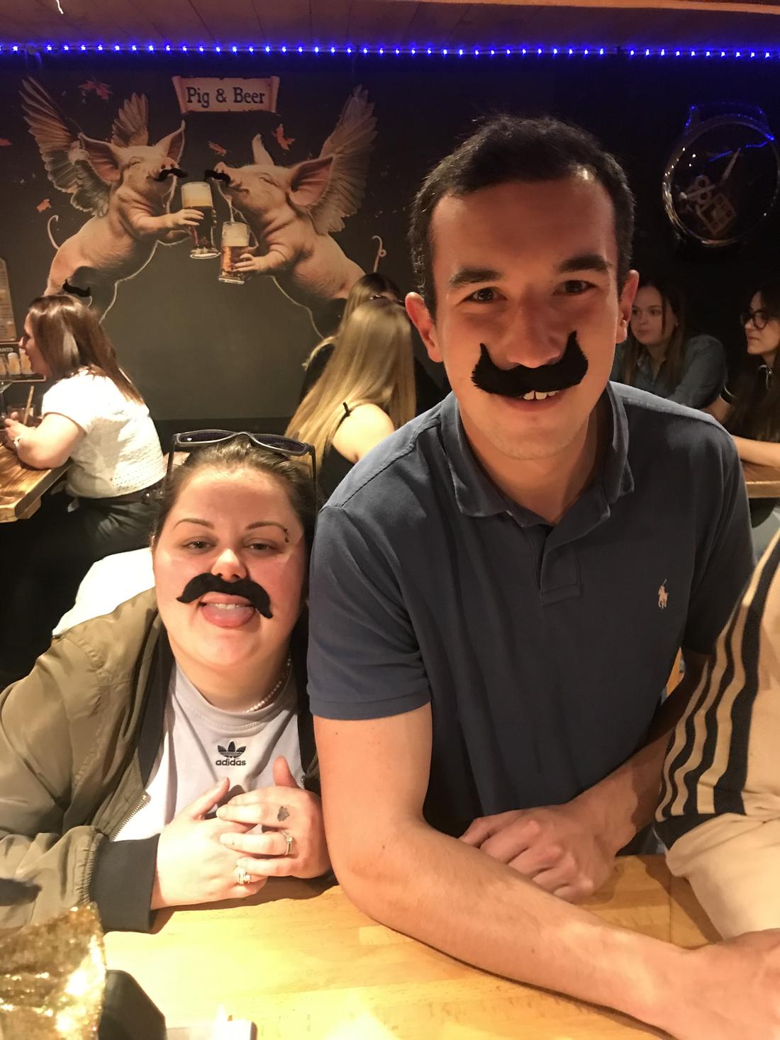 Deux clients avec de fausses moustaches devant la fresque des cochons ailés du PIC
